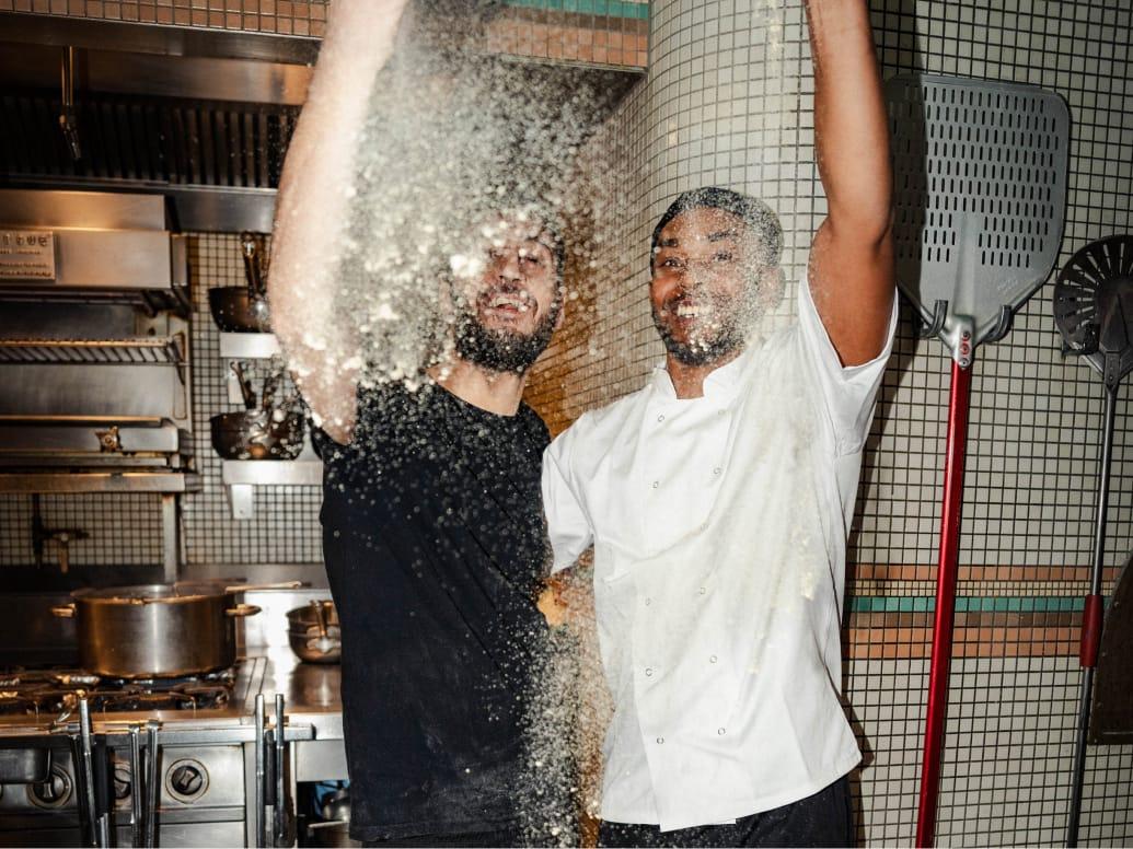 Image of two men throwing flour in a kitchen. On the left, the man is wearing a black t-shirt, on the right the man is wearing a Chef's uniform.