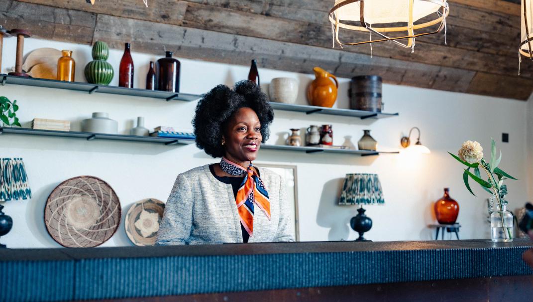 Image of a woman on the phone, standing behind the reception desk at Soho Farmhouse. She is wearing a grey jacket and has an orange and blue silk scarf around her neck. In the background, there are two shelves filled with ornaments.