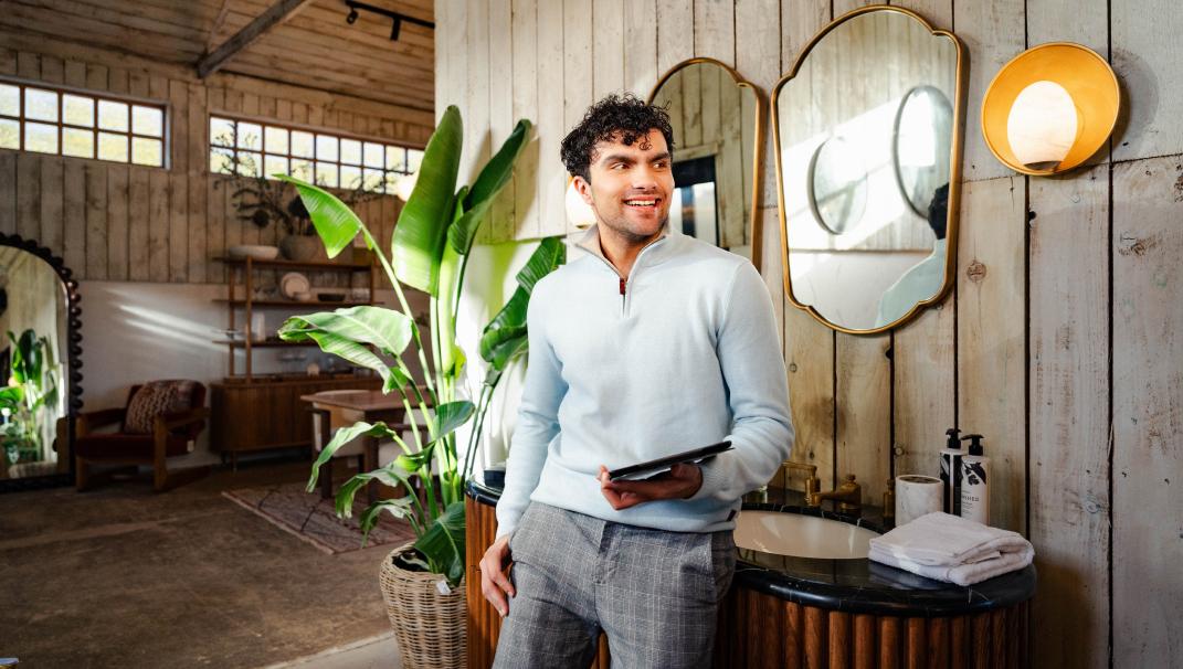 Mid shot of a man in the Soho Home retail space at Soho Farmhouse. The man is wearing a grey jumper and plaid trousers and is holding an iPad. Behind the smiling man is two mirrors over a red-tiled sink. To the left is an open rustic-styled bedroom.
