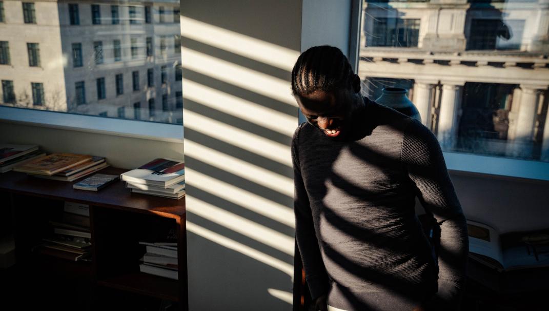 Image of a sunlit man standing in an office. The man is wearing a long-sleeved black top and black trousers and has rays of sunlight running across his face. His head is looking down.