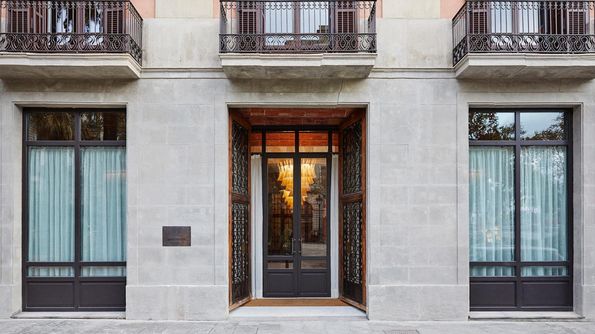 Photography image of a white stone Soho House building. The building features a central gold and dark brown front door, with two floor-to-ceiling brown double windows on either side. At the top of the image, you can see three banners.