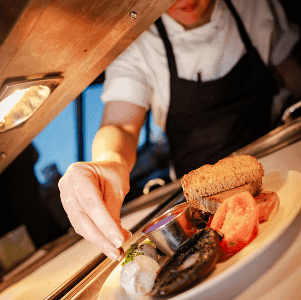 Image of a plated cooked breakfast that has been placed on the pass in the kitchen. A chef, wearing chef whites and an apron, is garnishing the poached eggs with greens.
