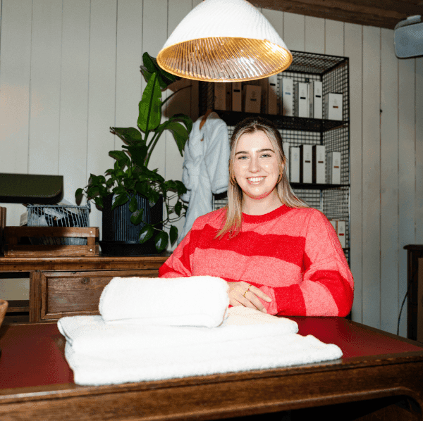 Image of a blonde host wearing a stripey pink and red jumper sat in the Soho Farmhouse spa. There are two folded white towels in front of her and a shelving unit with Cowshed products in the background.
