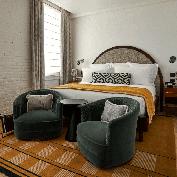 Image of a Soho House bedroom. The image featured an orange patterned carpet, velour green armchairs, a patterned rustic headboard and a double bed with white sheets and a yellow throw.