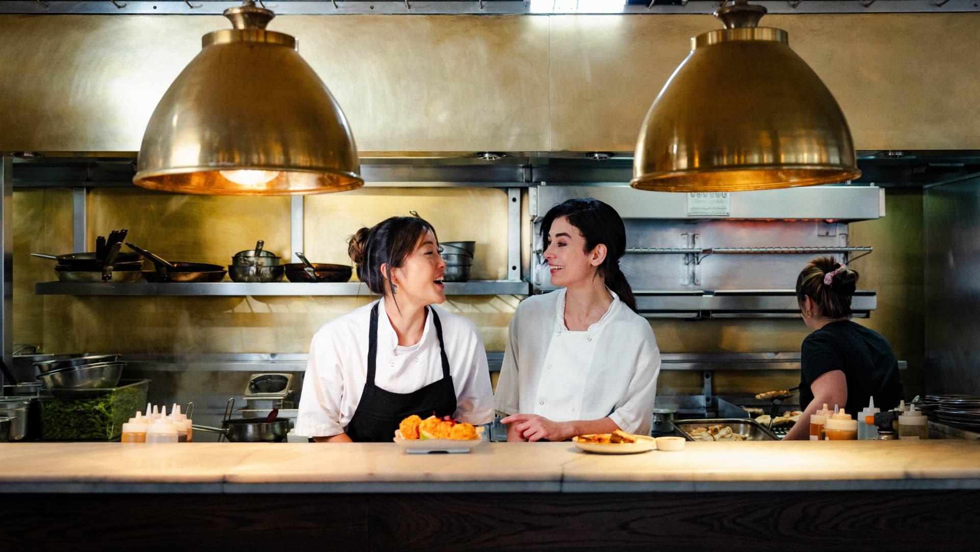 Image of two female chefs looking at one another laughing in the Shoreditch House kitchen. There are two large brass lights hanging from the ceiling. In front of them, there are two plated dishes ready to be served.