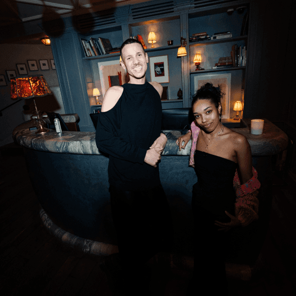 Image of two reception hosts standing in a dark-lit reception at Greek Street. The person on the left is a man wearing a black tank top and black jeans, he is smiling and looking to the left. The woman is positioned on the right, she is wearing a black bandeau top with a pink over-the-shoulder cardigan. The woman is also smiling.