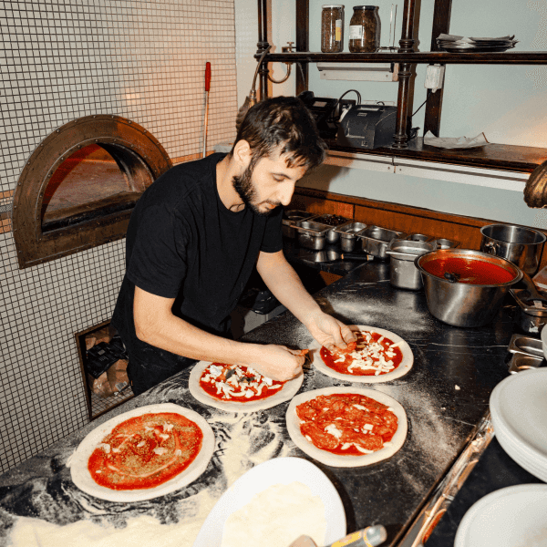 Image of a man wearing a black t-shirt making 4 pizza dishes on a dark stove counter. The image contains backlighting and is located in a kitchen with white tiles and green-painted walls at Cecconi’s.