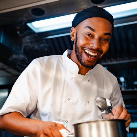 Close-up image of a smiling man wearing a white chef uniform holding a silver pan and serving spoon. The man is standing in a backlit kitchen at Dean Street Townhouse.