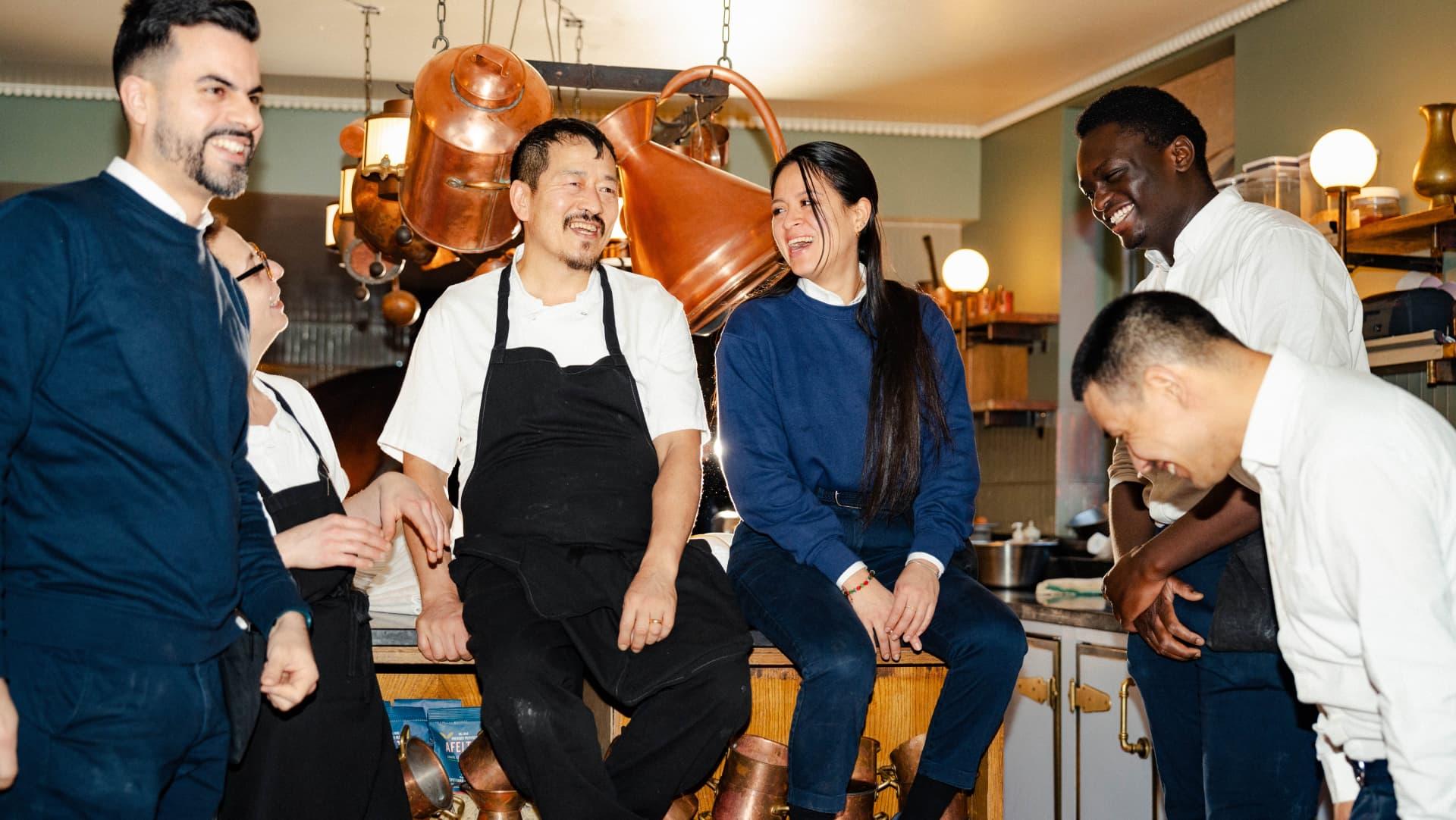 Image of a group of staff sat on the countertop of the kitchen at 40 Greek Street, Soho House. There is a combination of chefs, waiters, and bartenders in their uniforms. They are all laughing and smiling at one another.