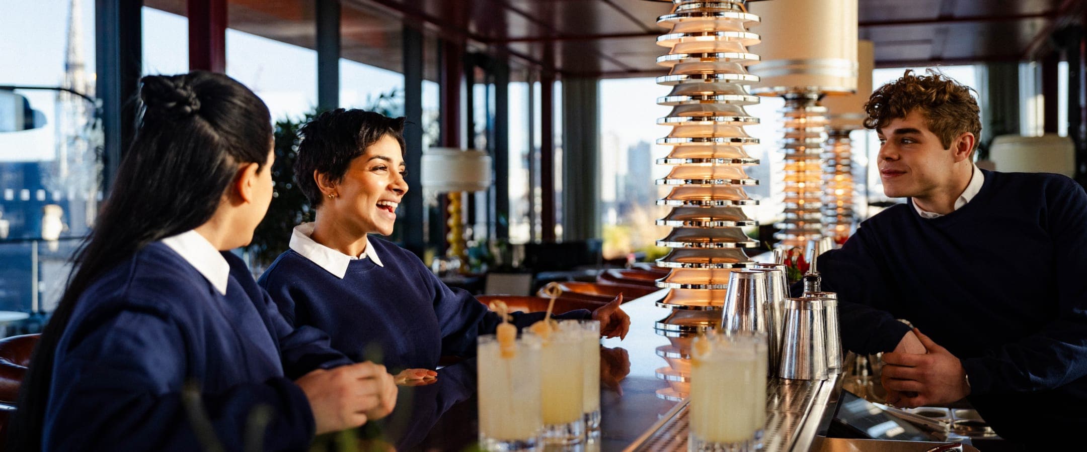 An image of three bar staff leaning against the top floor bar of 180 Strand. They are all wearing blue sweatshirts over white collared polos. On the bar there are five cocktails ready to be served.