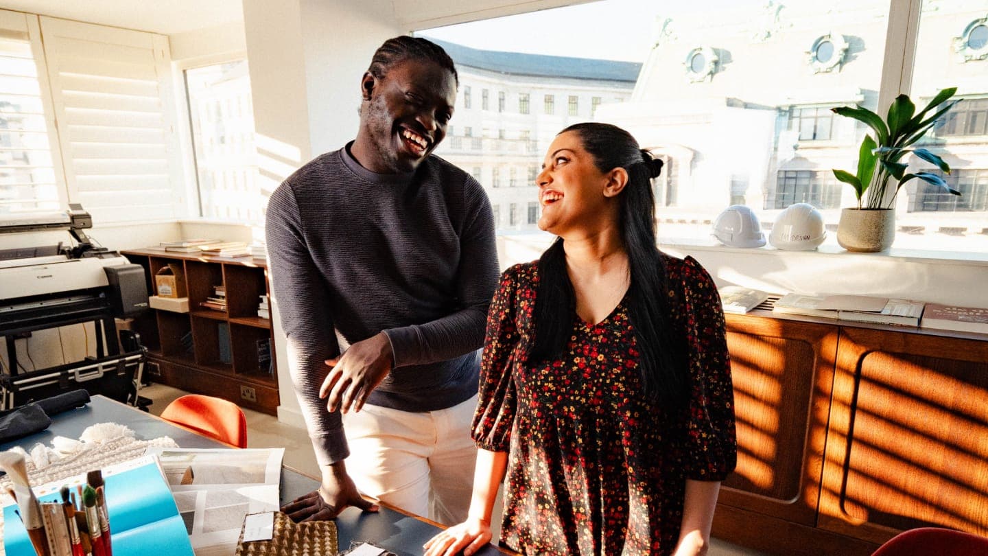 Image of two colleagues, one female and one male, are stood in front of a table at 180 Strand. There are fabric samples and design booklets in front of them on the table. They are looking at each other laughing.
