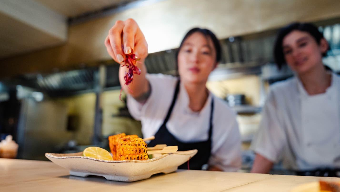 Close up image of a fried sweetcorn dish being plated in the pass of a kitchen. In the background, there are two female chefs in chef whites.