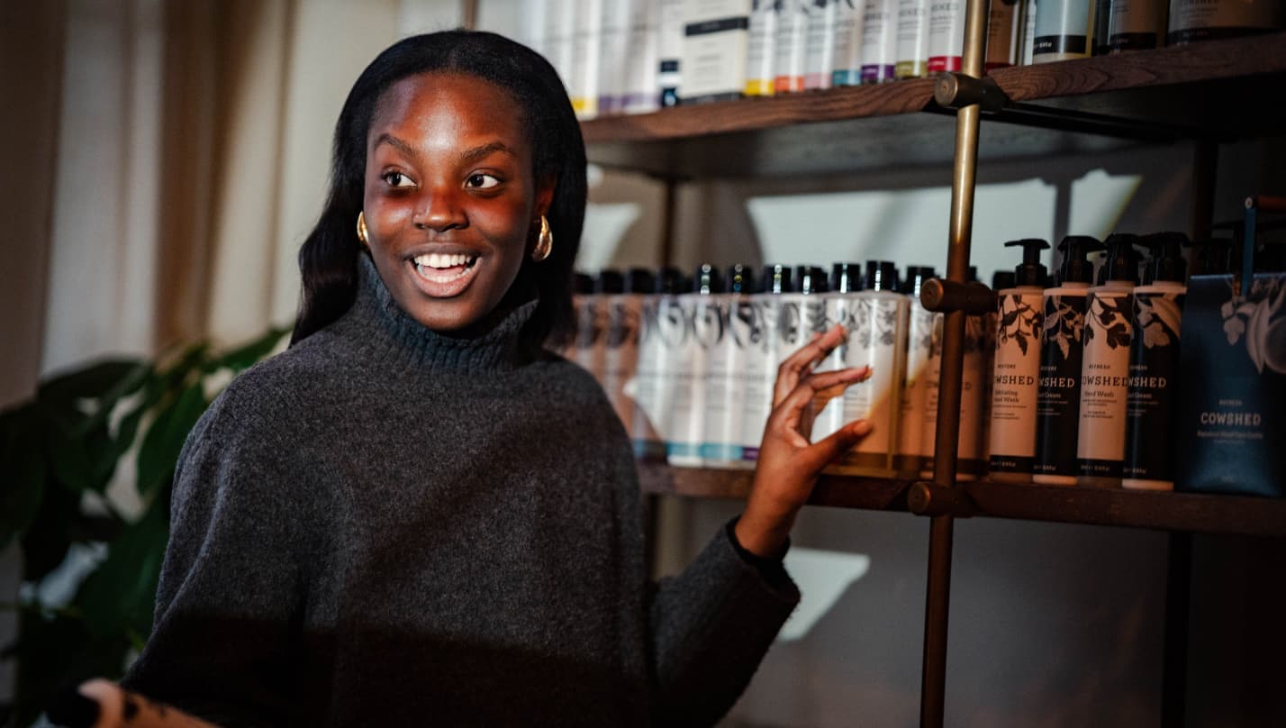 Image of a spa host in front of a display of Cowshed products. She is holding a bottle of hand cream in one hand, describing it to others around her.