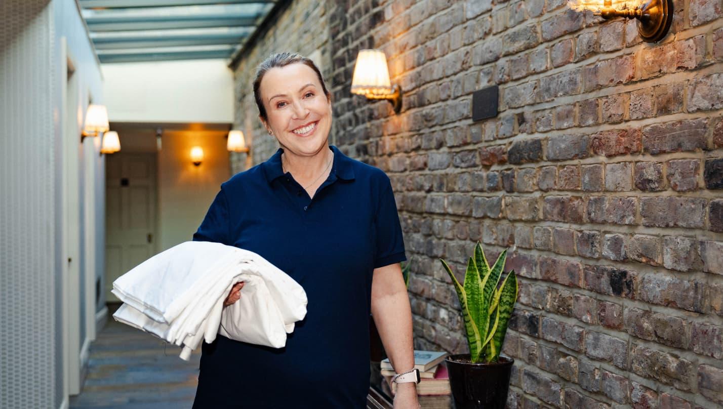 Image of a cleaner in the corridor of Kettner's hotel. She is carrying a pile of white towels and smiling. The background is an exposed brick wall, with a table of books and a plant.