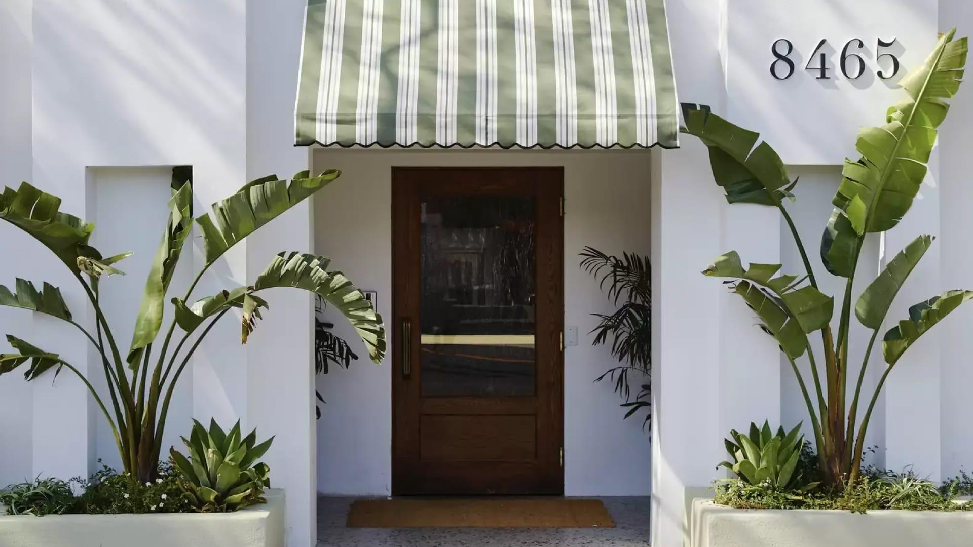 Photography of LA's Soho House Holloway. The image depicts a white building with a central brown front door. On either side of the door are two green palm trees, and the top of the door features a green and white door cover.
