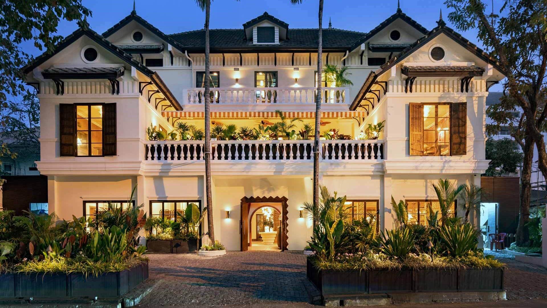 Image of a tropical Soho House home. The image features a long shot of an entirely white building, with wide open wooden windows. There is a yellow light which is visible through the open windows and lots of palm trees and green flowers growing on the balcony and in the front garden.