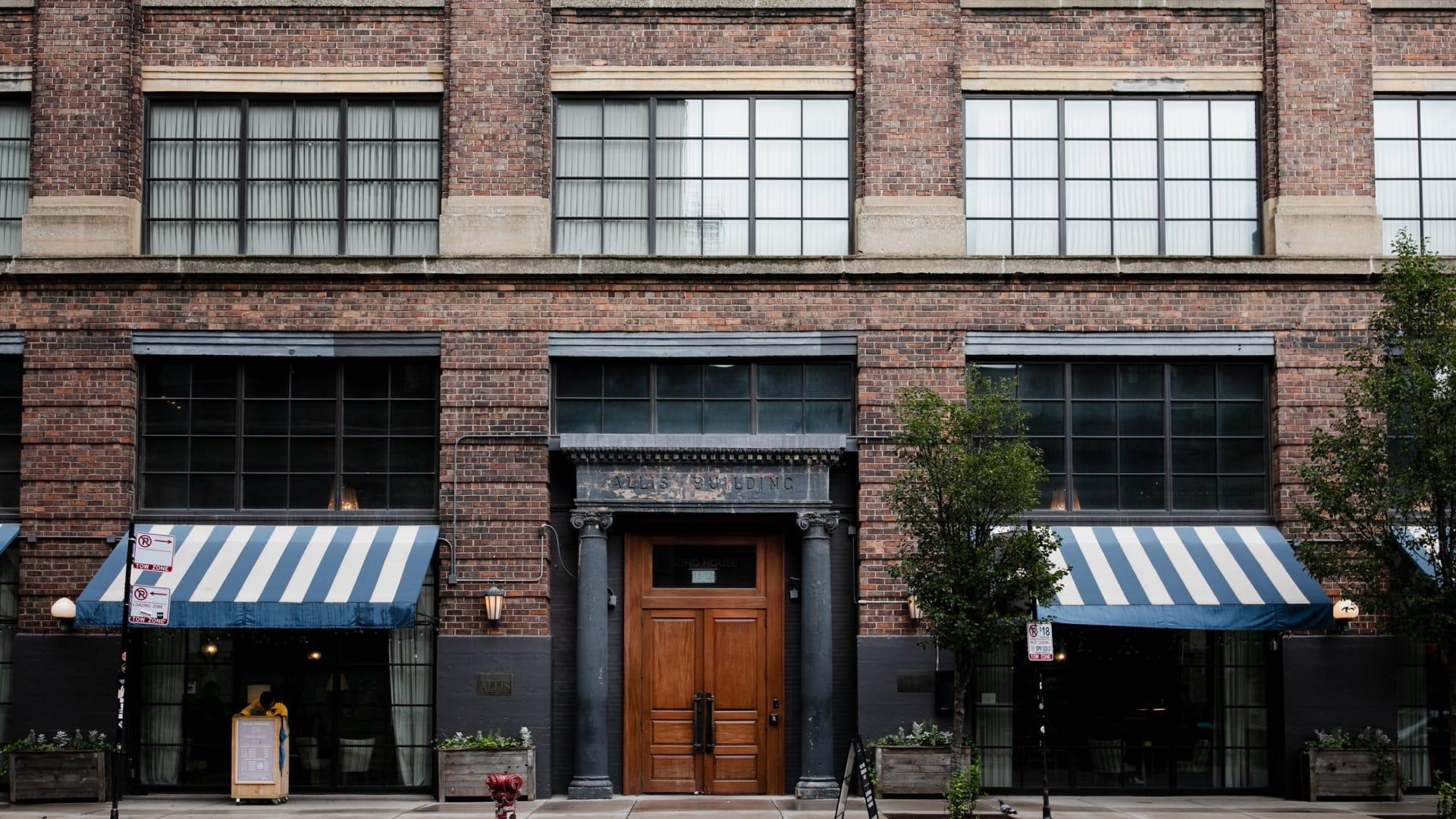 Mid shot of a brown bricked Soho house building. The focal point of the image features a brown wooden oak door with navy blue rain-washed pillars. On either side of the door is a double window, with white and navy stripped sun covers.