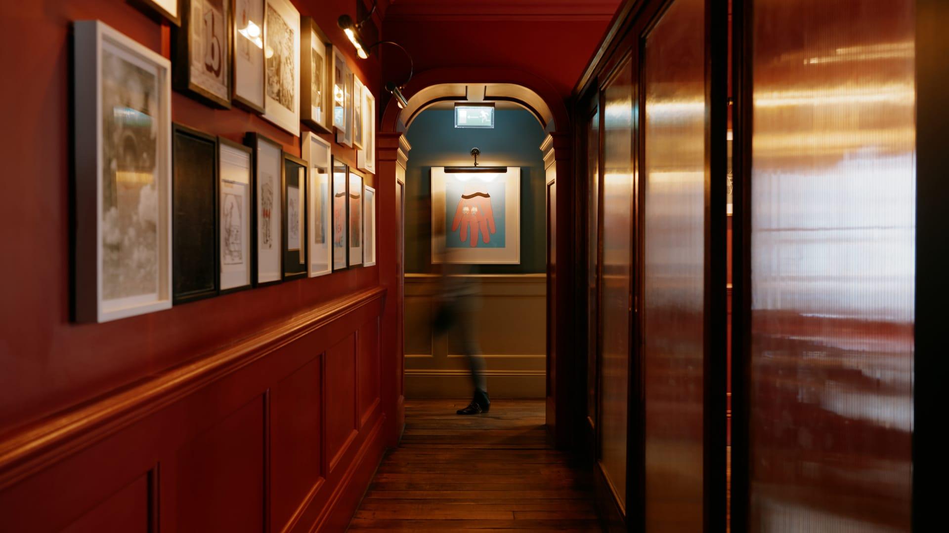 Focused image of a red and ebony wooden hall way. On the left-hand side is a wall with a series of black and white photo-frames. The images are not in focus. At the end of the hall is an unfocused person walking.