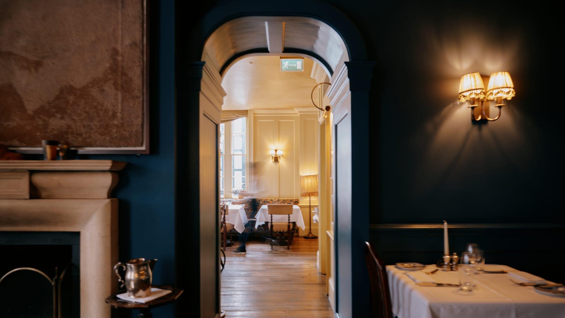 Photograph of an editorial-styled shot. The image features an internal hall archway in rich navy, with a set table featuring a white tablecloth and silverware on the right. Through the doorway you can see a mustard armchair and a small table with a white tablecloth.