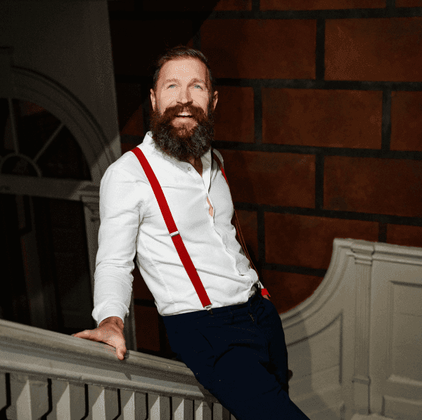 Image of a smiling man with a beard stood on a white stair case with a red brick wall. The man is wearing a white shirt with red braces and clack trousers.