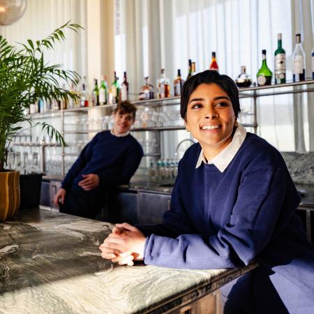Focused image of a woman in a blue jumper with a white shirt leaning against a countertop. There is a man in the background wearing a black jumper. Both people are staging in the Soho Farmhouse bar