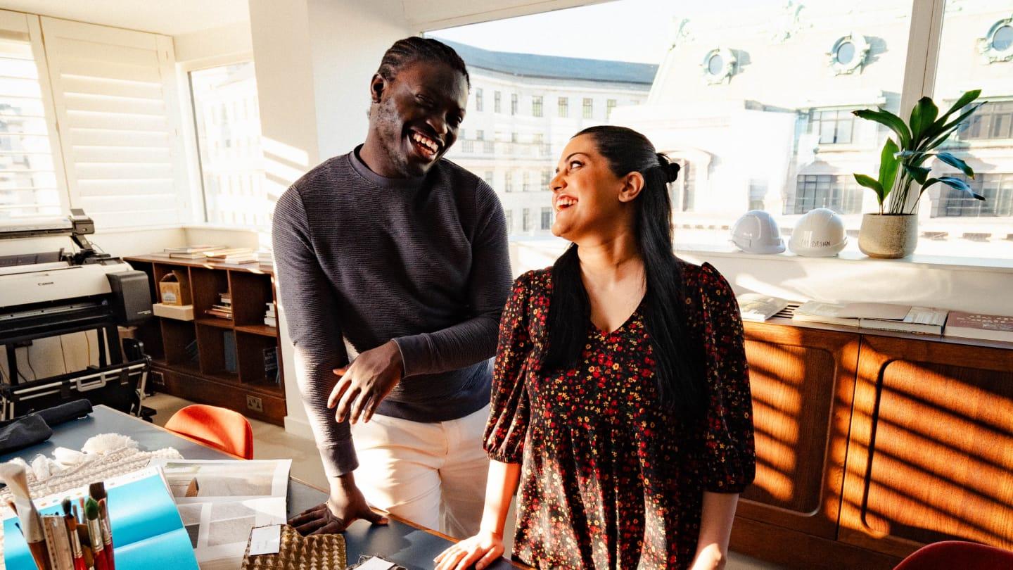 Two multi-cultural people stood in a Soho House office, smiling at each other. The man on the left is wearing a navy jumper. The women positioned on the right is wearing a floral dress
