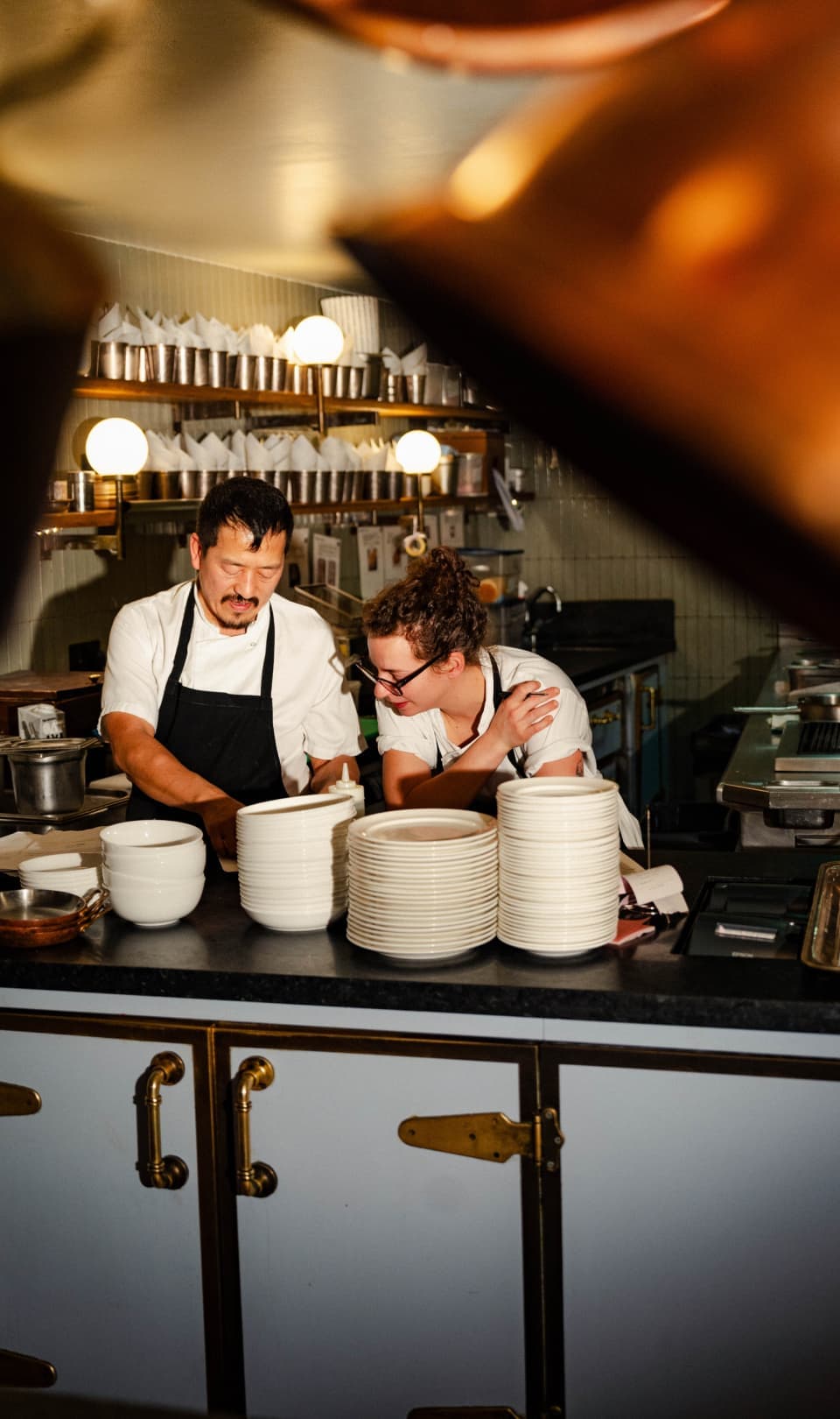 Two chefs wearing aprons working at a kitchen counter with stacked plates, warm lighting, and copper pots hanging overhead
