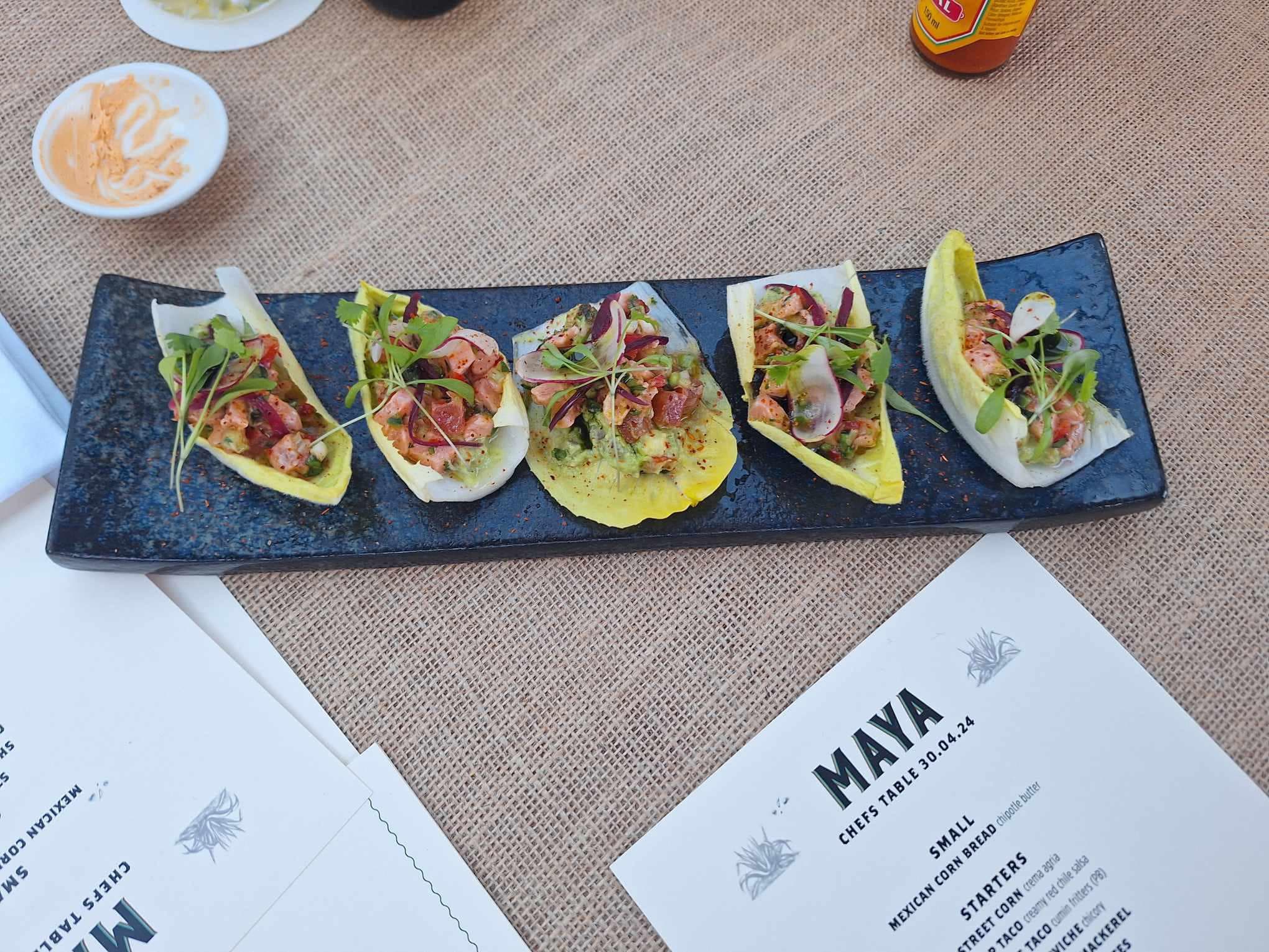 Five endive leaves filled with diced vegetables, herbs, and microgreens on a dark rectangular plate, served on a burlap-covered table with a "MAYA" menu and sauce on the side